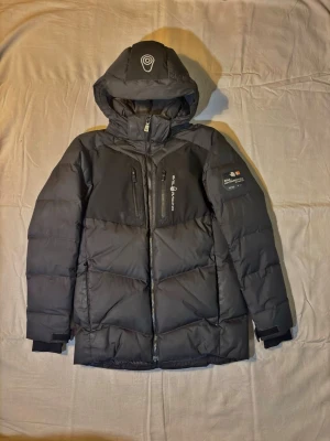 Svart dunjacka från sail racing - Snygg svart dunjacka från Sail racing med huva och dragkedja framtill. Jackan har flera fickor med dragkedja, logga på bröstet och ärmen samt en modern, vadderad design som håller dig varm under kalla dagar.