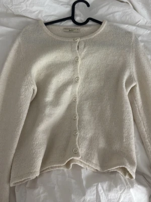 Krämvit kofta från Femme Select - Mysig krämvit kofta från Femme Select med rund halsringning och raka långa ärmar. Koftan har en rak passform och stängs med sju matchande knappar framtill. Perfekt att slänga över en topp för en enkel och stilren look.