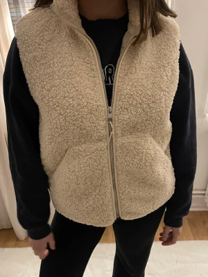 Beige teddyväst med dragkedja - Mysig beige teddyväst med stora fickor framtill. Tillverkad i fluffigt teddy-material som ger en trendig och avslappnad look. Perfekt passform för både över en hoodie eller tshirt. Fodrad insida och hög krage för extra komfort. Använd 1 gång