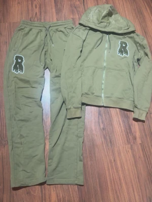 Olivgrön loungewear-set med hoodie risky - Snyggt olivgrönt loungewear-set med matchande hoodie och byxor. Båda plaggen har en cool patch med ett droppande 'R' på bröstet och benet. Hoodien har dragkedja och fickor, byxorna har dragsko i midjan. Perfekt för chill dagar hemma.