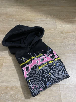 Svart hoodie med spindelnäts-tryck - Säljer en svart hoodie med huva och dragsko. Framsidan har ett stort tryck med spindelnät, gula stjärnor och texten 'nevermind the spider, here’s the slime' i gult och rosa. Materialet känns mjukt och bekvämt, perfekt för en avslappnad streetwear-look.