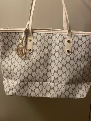 Vit shoppingväska med märket M - Snygg shoppingväska lik Michael Kors i vitt med grått M-mönster över hela väskan. Väskan har två handtag, guldfärgade detaljer och en rund M hänge framtill. Tillverkad i tåligt konstläder och har rymligt innerfack med flera mindre fack.