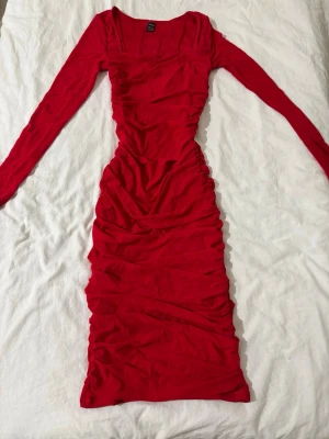 Röd långärmad klänning från SHEIN - Trendig röd klänning från SHEIN med långa ärmar och figurnära passform. Klänningen har rynkade detaljer längs sidorna och en fyrkantig ringning. Perfekt för dig som vill sticka ut med en färgstark look.