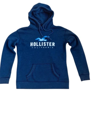 Blå hoodie från Hollister - Snygg blå hoodie från Hollister med vit logga och blå fågeltryck på bröstet. Klassisk känguruficka framtill och justerbar dragsko i huvan. Perfekt för en avslappnad och cool stil. Mjuk bomullsblandning som känns skön mot huden.