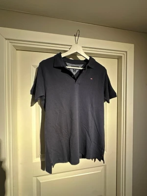 Mörkblå piké från Tommy Hilfiger - Klassisk mörkblå pikétröja från Tommy Hilfiger med liten logga på bröstet. Skjortan har korta ärmar, krage och knappar vid halsen. Tillverkad i mjuk bomull som känns skön mot huden. Perfekt för en clean och stilren look.