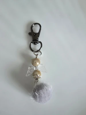Nyckelring med ängel och fluffboll - Söt nyckelring med en ängel gjord av pärlor i guldton och transparenta vingar, samt en mjuk ljusgrå fluffboll. Metallfäste i silverfärg gör den enkel att sätta på väskan eller nyckelknippan. Perfekt accessoar för att ge lite extra stil.