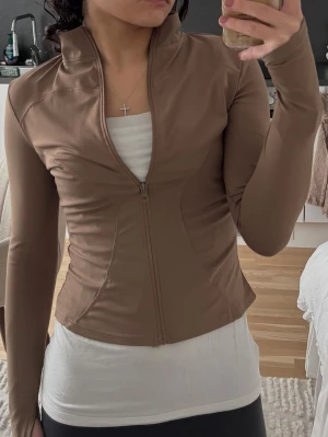 Brun sportjacka med dragkedja - Snygg brun sportjacka med figurnära passform och hel dragkedja framtill. Jackan har hög krage och långa ärmar med tumhål. Tillverkad i ett stretchigt, mjukt material som passar perfekt till träning eller som ett extra lager. .