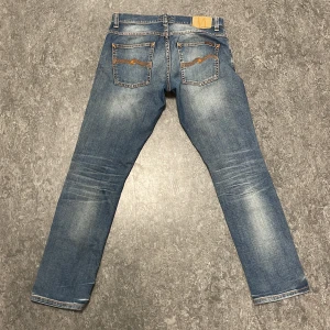 Blå Nudie Jeans - Säljer ett par klassiska blå jeans från Nudie Jeans med snygga slitningar och kontrastsömmar. Modellen har raka ben, fem fickor och läderpatch bak i midjan. Perfekta för dig som gillar en avslappnad men trendig look.