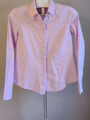 Ljusrosa skjorta från Park Lane - Klassisk ljusrosa skjorta från Park Lane med lång ärm och knappar framtill. Skjortan har en stilren krage och diskret broderad logga på bröstet. Perfekt för en clean och fräsch look, lätt att matcha med jeans eller kjol.