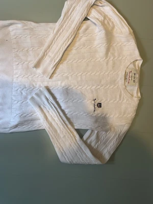 Vit kabelstickad tröja från Bondelid - Kabelstickad vit tröja från Bondelid med rund halsringning och broderad logga på bröstet. Tröjan har långa ärmar och ribbstickade muddar vid ärmslut och nederkant. Klassisk och stilren design i mjukt material.