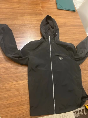 Svart vindjacka från Prada - Snygg svart vindjacka från Prada med huva och dragkedja framtill. Jackan har en minimalistisk design med Pradas ikoniska trekantslogga på bröstet. Tillverkad i lätt och tåligt material som står emot vind. Perfekt för dig som vill ha en stilren och exklusiv look.