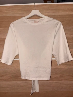 Vit topp med knyt från Nelly - En vit topp från Nelly med trekvartsärm och öppen rygg med knytband vid ryggen. Toppen har rund halsringning och är gjord mjuk bomull. Aldrig använd - utan prislapp.