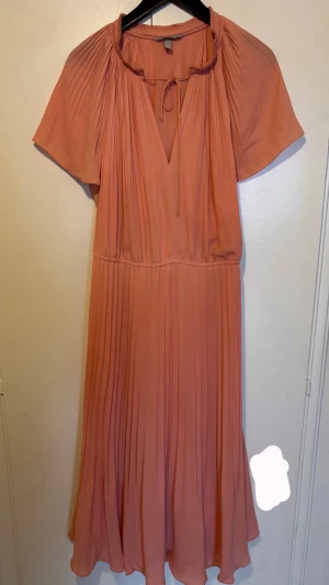 Lång klänning  - Säljer en persikofärgad kortklänning från H&M med plisserad kjol, v-ringning och knytband vid halsen. Klänningen har korta ärmar.