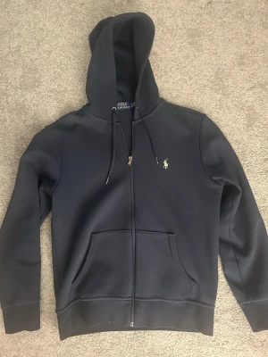 Mörkblå zip hoodie från Polo Ralph Lauren - Snygg mörkblå hoodie med dragkedja från Polo Ralph Lauren. Klassisk design med huva, dragsnören och känguruficka framtill. Liten broderad logga på bröstet. Perfekt för en avslappnad och stilren look.