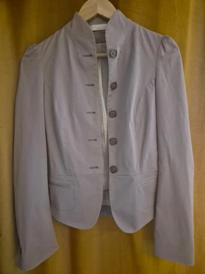 Ljusgrå kavaj med ståkrage - Snygg ljusgrå kavaj med ståkrage och fem knappar framtill. Kavajen har figurnära passform, lätt puffade axlar och två fickor framtill. Perfekt för en stilren och modern look. 