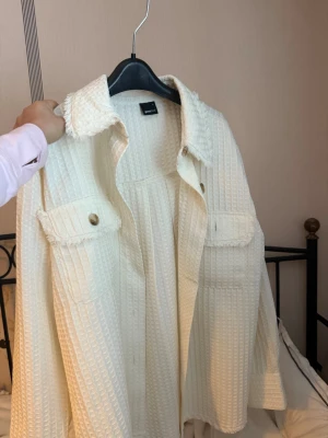 Vit och en brun overshirt från Gina Tricot - Stilren vit overshirt från Gina Tricot i strukturerat tyg med rutmönster. Skjortan har två stora bröstfickor med knappar och råa kanter på kragen för en trendig look. Perfekt att bära öppen över en t-shirt eller stängd som jacka.