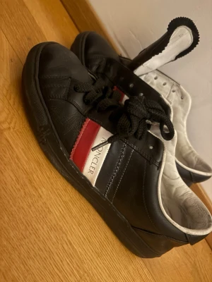 Moncler sneakers svart röd vit äkta - Snygga sneakers från Moncler i svart läder med klassiska ränder i rött, vitt och blått på sidan. Skorna har svarta skosnören, vit insida och Moncler-logga på plösen. Perfekta för dig som gillar stilrena och sportiga skor.