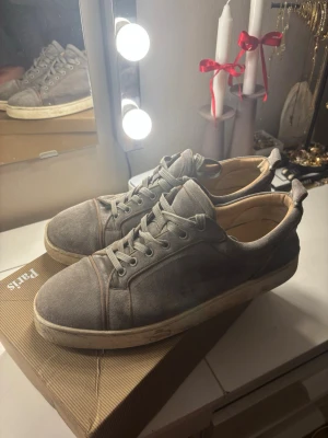 Gråa Christian Louboutin sneakers - Snygga gråa sneakers från Christian Louboutin med klassisk röd sula och lågt skaft. Skorna är tillverkade i mocka och har vita snören samt diskreta sömdetaljer. Perfekta för dig som vill ha en exklusiv touch på din outfit. De är i använt skick som ni ser på bilderna. Ingår dustbag och kvitton och certifikat