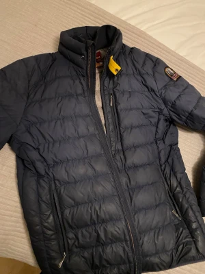 Mörkblå dunjacka från Parajumpers helt äkta - Snygg mörkblå dunjacka från Parajumpers med gul detalj vid kragen och patch på ärmen. Jackan har två dragkedjeförsedda fickor framtill och en innerficka. Lätt och smidig modell med quiltat mönster och hög krage.