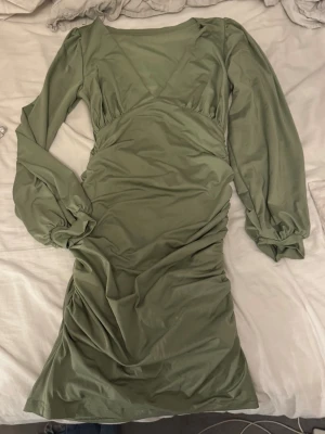 Olivgrön långärmad klänning - Snygg olivgrön klänning med lång ärm och v-ringad hals. Klänningen har en figurnära passform och lätt rynkade detaljer längs sidorna. Tillverkad i mjukt material som känns bekvämt mot huden. Perfekt för dig som gillar stilrena plagg med en twist.