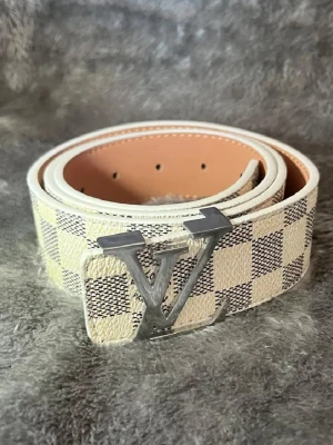 Snygg Rutig beige bälte - Snyggt bälte från i beige och vitt med klassiskt rutigt mönster och stor logga i silver på spännet. Bältet har exklusiv känsla och passar perfekt till jeans eller byxor för en trendig look.