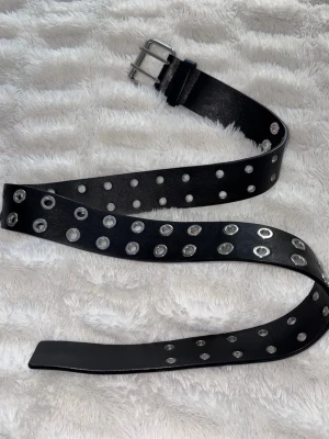 Svart bälte med nitar och hål - Svart bälte med dubbla rader av silvriga metallhål och nitar längs hela bandet. Klassisk metallspänne framtill. Perfekt accessoar för att ge en edgy touch till din outfit. Passar om man har typ mindre än storlek 26 i byxor. Minns inte märke men typ monki eller något 