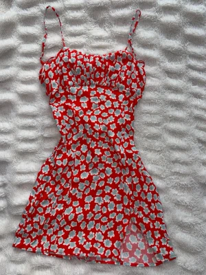 Röd blommig kortklänning med knyt  - Söt röd kortklänning med vita och ljusblå blommor, tunna axelband och knyt bak och dragkedja. Klänningen har en figurnära passform och är gjord i ett lätt, luftigt material som passar perfekt till varma dagar. Perfekt för dig som gillar färgglada och lekfulla plagg. Perfekt till skolavslutning. Storlek S och knappt använd