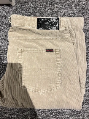 Beiga manchesterbyxor från Edwin - Snygga beiga manchesterbyxor från Edwin med klassisk femficksdesign och smalspårig manchester. Byxorna har en svart patch bak i midjan och en liten Edwin-logga på bakfickan. Perfekta för dig som gillar retrostil och vill ha något unikt i garderoben.