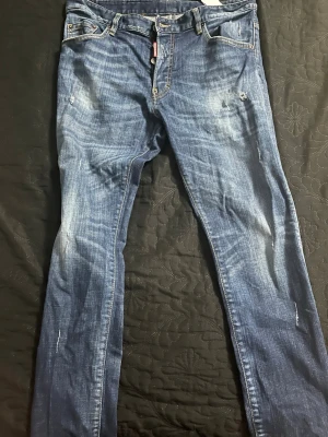 Blå jeans med slitningar och rosdetalj - Säljer ett par blå jeans från Dsquared2 med snygga slitningar och tvättade detaljer. Baktill finns en patch med rosor som ger en unik touch. Klassisk femficksmodell och smal passform. Perfekta för dig som gillar streetstyle och vill sticka ut.