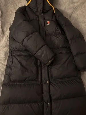 Svart Fjällräven Expedition dunjacka XL - Säljer en svart lång dunjacka från Fjällräven Expedition Series, dammodell i storlek XL. Jackan har gula detaljer vid huvan, två stora framfickor med lock och tryckknappar samt klassisk Fjällräven-logga på bröstet. Perfekt för kalla vinterdagar. Inga defekter alls som ny.