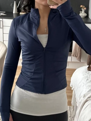 Mörkblå sportjacka med dragkedja - Snygg mörkblå sportjacka med hel dragkedja framtill och hög krage. Figurnära passform och långa ärmar med tumhål. Tillverkad i ett stretchigt, mjukt material som passar perfekt till träning eller promenader.