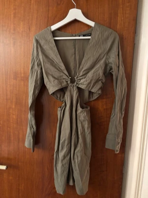 Olivgrön omlottklänning med cut-out - Trendig olivgrön kortklänning från Zara med lång ärm, djup v-ringning och snygg cut-out framtill. Klänningen har omlottdetalj och knyte vid midjan som ger en cool och modern look. M men mer som en S