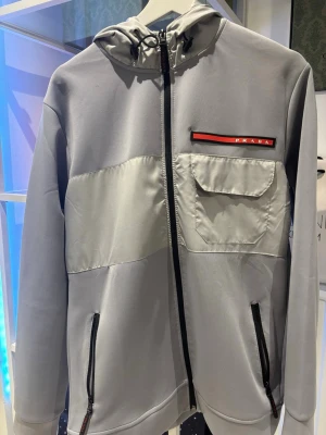 Grå vindjacka med huva från Prada - Tjena, säljer min tvärfeta Prada cardigan/overshirt. Ja kan är i toppskick förutom en liten skada vid loggan på bilden men annars topp skick. Skriv för fler bilder eller frågor!