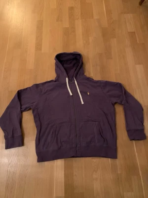 Ralph Lauren  - Säljer en lila hoodie från Polo Ralph Lauren med dragkedja framtill och vita snören i huvan. Klassisk liten logga broderad på bröstet. Hoodien har två fickor fram och ribbade muddar vid ärmar och nederkant. Perfekt för en chill och avslappnad stil.