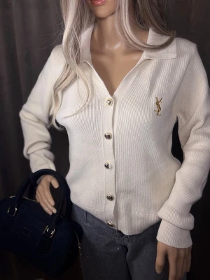 Vit kofta från Yves Saint Laurent - Stilren vit kofta från Yves Saint Laurent med guldiga knappar och broderad YSL-logga på bröstet. Koftan har en klassisk krage, ribbstickade muddar och är tillverkad i ett mjukt, stickat material. Perfekt för en trendig och elegant look.