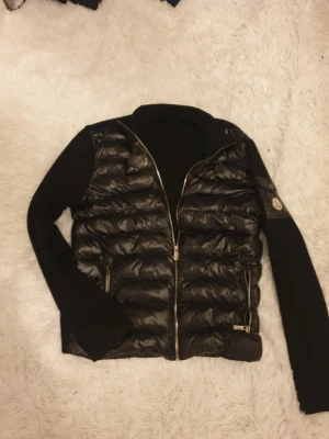 Svart dunväst från Moncler - Snygg svart dunväst från Moncler med quiltad front och två dragkedjefickor. Ärmar och rygg i mjukt tyg ger en cool kontrast till den glansiga dunfronten. Klassisk Moncler-logga på ärmen och dragkedja framtill. Perfekt för lager-på-lager och streetstyle. Storlek M men passar också S. Skinnet har förstörts lite närmare du kollar så syns det. Kolla sista bilden