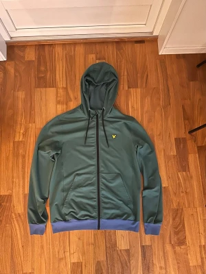Grön zip från Lyle & Scott - Snygg grön hoodie med dragkedja från Lyle & Scott. Har huva med dragsko, två fickor framtill och ribbade muddar i blått vid ärmslut och nederkant. Liten gul logga på bröstet. Perfekt för en chill och stilren look.