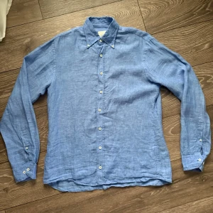 Blå linneskjorta  - Snygg blå skjorta i linne från Oscar Jacobson med klassisk krage och knappar hela vägen framtill. Skjortan har långa ärmar med manschett och en avslappnad passform. Perfekt för en stilren och chill look.