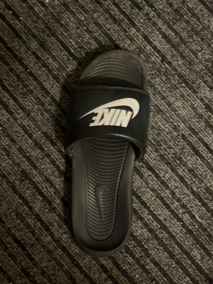 Svart Nike badsandal - Klassisk svart badsandal från Nike med vitt logotryck på remmen. Sandalen har en bred rem över foten och räfflad sula för extra grepp. Tillverkad i syntetmaterial och perfekt för chill dagar eller efter träningen.