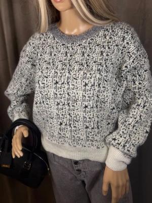 Gråmelerad stickad tröja med mönster - Säljer en gråmelerad stickad tröja med svart och vitt abstrakt mönster. Tröjan har rund halsringning, ribbade muddar och nederkant samt en avslappnad passform. Perfekt för dig som gillar en stilren och mysig look.
