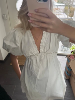 Vit kort puffärmad klänning - Söt vit kortklänning med puffärmar och djup v-ringning. Klänningen har volangdetaljer längs urringningen och en markerad midja som ger en feminin siluett. Tillverkad i bomull för en luftig och bekväm känsla.