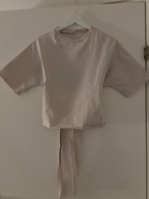 Ljusrosa  topp med knyt - Trendig ljusrosa croppad topp med korta ärmar och knytband i midjan som kan knytas bak. Perfekt för en avslappnad och snygg look. Tillverkad i mjuk bomull för extra komfort.