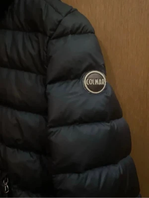 Mörkblå dunjacka från Colmar - Snygg mörkblå dunjacka från Colmar med glansig finish och klassisk puffer-design. Jackan har huva, dragkedja framtill och två sidofickor. Colmar-logga på ärmen. Det är ett väldigt litet hål på armen men ingenting som märks skriv om man vill ha bild så skickar jag!💛