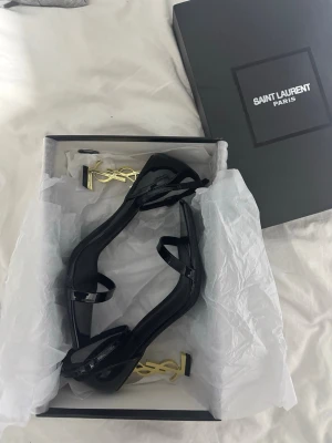 YSL klackar - Säljer ett par svarta lackade pumps från Saint Laurent med öppen tå, smala remmar och ikonisk guldig YSL-logga som klack. Snygg fyrkantig tå och justerbar ankelrem. Perfekta för dig som vill sticka ut med lyxig design och cool detalj.