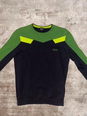 Grön och svart tröja BOSS -  Tröja från Hugo Boss i svart och grönt med neongula detaljer vid axlarna. Rund halsringning med ribbstickad kant och diskret BOSS-logga på bröstet. Perfekt för dig som gillar sportig och modern stil.