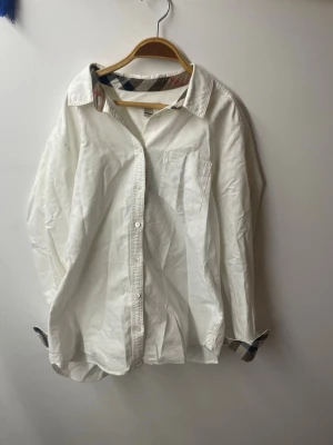 Vit skjorta från Burberry Brit - Klassisk vit skjorta från Burberry Brit i bomull med lång ärm och knappar framtill. Ikoniskt rutigt mönster syns på insidan av kragen och manschetterna. Perfekt för en stilren och tidlös look.