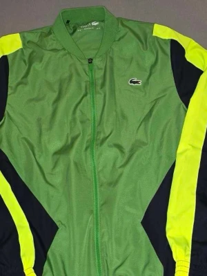 Grön vindjacka från Lacoste - Den är aldrig använd den är bara tvättad. Så helt ny är den. Säljer billigt pågrund av att den blev för liten på mig. Har M nu. Hör av er vid frågor! Sportig grön vindjacka från Lacoste med dragkedja framtill. Jackan har svarta och neongula detaljer längs ärmarna och axlarna samt Lacoste-logga på bröstet och ryggen. Perfekt för en streetwear-look. Helt ny 