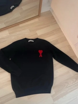 Svart stickad tröja från Ami Paris - Svart stickad tröja från Ami Paris med röd hjärtlogga och bokstaven A broderad på bröstet. Tröjan har rund halsringning och långa ärmar. Perfekt för dig som gillar stilrena plagg med en twist.