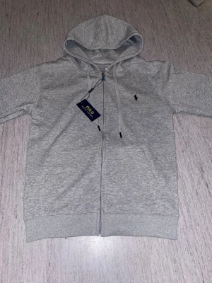 Grå hoodie från Polo Ralph Lauren - Klassisk grå zip-hoodie från Polo Ralph Lauren med huva och dragsko. Broderad logga på bröstet och två fickor framtill. Tillverkad i mjuk bomullsblandning