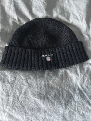 Svart ribbstickad mössa från GANT - Svart ribbstickad mössa från GANT med broderad logotyp och sköld framtill. Klassisk modell med uppvikt kant och mjukt material som håller värmen. Perfekt accessoar för kyliga dagar.